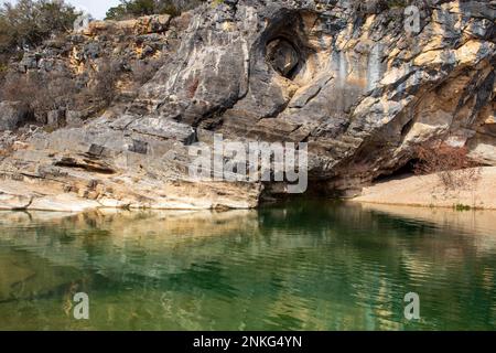 Eine Wand aus geologischen Kalkstein- und Granitfelsformationen bildet eine Höhle und ein Loch in der Wand mit Reflexionen im grünen Teich in Pedernales Falls Stockfoto