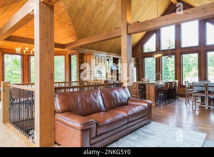 Dunkelbraunes Ledersofa vor der Treppe im Wohnzimmer plus Küche mit Insel- und Frühstückstisch im gemahlenen östlichen weißen Kiefernholzhaus. Stockfoto