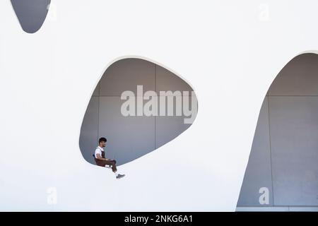 Junger Mann mit Laptop, der an der Wand sitzt Stockfoto