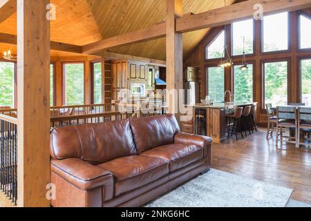 Dunkelbraunes Ledersofa vor der Treppe im Wohnzimmer plus Küche mit Insel- und Frühstückstisch im gemahlenen östlichen weißen Kiefernholzhaus. Stockfoto