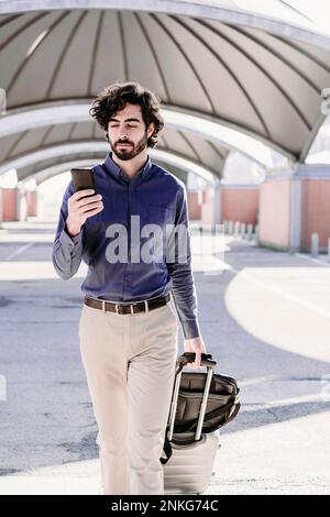 Ein Geschäftsmann, der mit Gepäck unterwegs ist und ein Smartphone benutzt Stockfoto