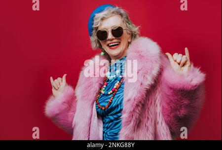 Glückliche Seniorin in rosa Pelzmantel, gestrickt vor rotem Hintergrund Stockfoto