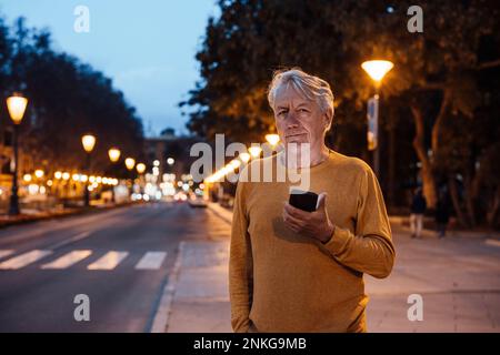 Lächelnder Senior-Mann, der mit dem Handy auf dem Bürgersteig stand Stockfoto