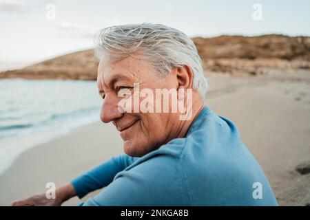 Lächelnder Senior-Mann, der bei Sonnenuntergang am Strand saß Stockfoto