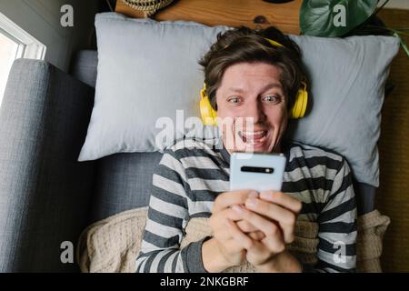 Aufgeregter Mann mit Smartphone und Kopfhörern, der zu Hause auf dem Bett liegt Stockfoto