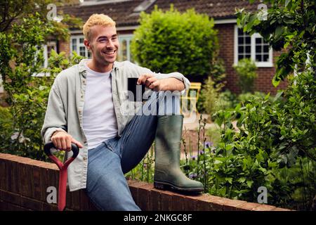 Glücklicher junger Mann mit Gummistiefel, der auf dem Zaun im Garten sitzt Stockfoto