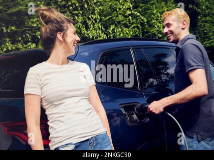 Glückliche Frau, die mit ihrem Freund spricht, der ein Elektroauto auflädt Stockfoto