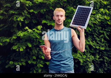 Junger Mann lädt Smartphone über Solarpaneel vor Pflanzen Stockfoto