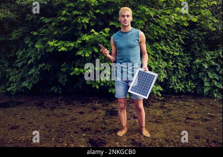 Junger Mann, der im Fluss steht und ein Handy hält, das von einem Solarmodul aufgeladen wird Stockfoto