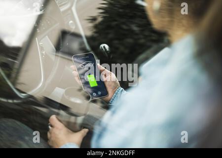 Frau mit Thermosflasche, die die App zum Laden von Elektroautos verwendet, durch Glas gesehen Stockfoto