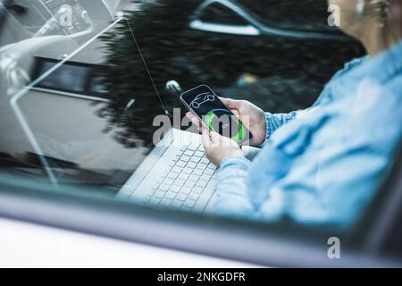 Reife Frau mit Laptop- und Elektroauto-Lade-App auf dem mobilen Bildschirm, durch Glas gesehen Stockfoto