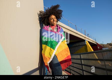 Lächelnde junge Frau, eingewickelt in Regenbogenfahne, die sich an die Wand lehnt Stockfoto