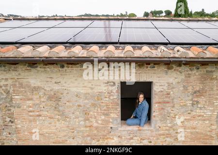 Lächelnde Frau, die auf dem Fensterbrett sitzt, mit Sonnenkollektoren auf dem Dach Stockfoto