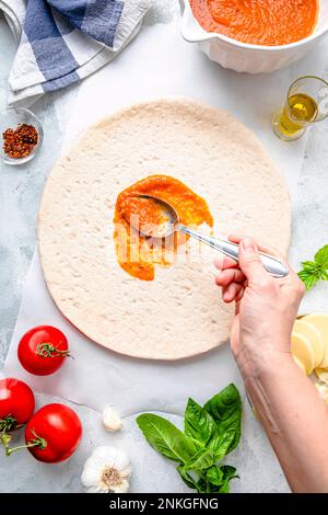 Hand einer reifen Frau, die Tomatensauce mit Löffel auf Pizzateig verteilt Stockfoto