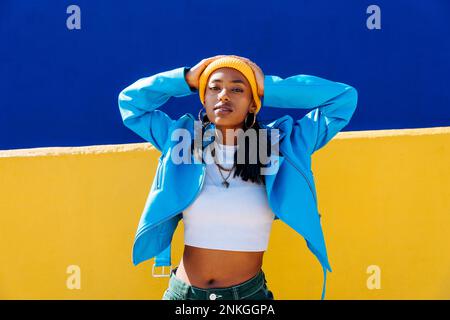 Selbstbewusste junge Frau in blauer Jacke, die vor einer gelben Wand steht Stockfoto