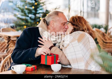 Ein älterer Mann mit Geschenkbox küsst eine ältere Frau im Café Stockfoto