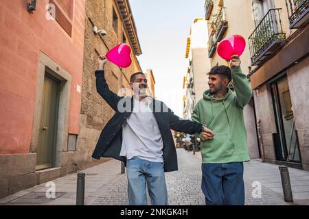 Glückliches schwules Paar, das Spaß mit roten herzförmigen Ballons in den Gebäuden der Straße hat Stockfoto