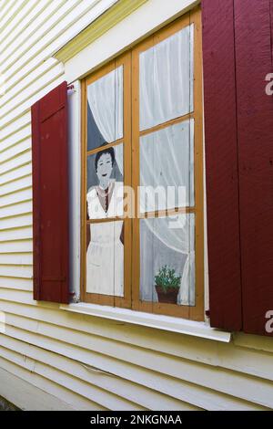 Frau steht in einer gefälschten bemalten Fensterszene mit roten hölzernen Fensterläden an der äußeren Seitenwand des alten, um 1840 Jahre alten, weißen horizontalen Holzplanken-Hauses. Stockfoto