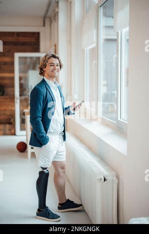 Glücklicher junger Geschäftsmann mit Prothesen, der am Fenster steht Stockfoto
