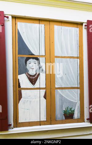 Frau steht in einer gefälschten bemalten Fensterszene mit roten hölzernen Fensterläden an der äußeren Seitenwand des alten, um 1840 Jahre alten, weißen horizontalen Holzplanken-Hauses. Stockfoto