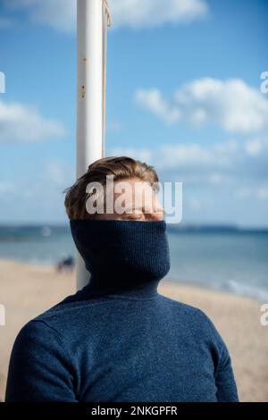 Ein Mann, der sein Gesicht mit Rollkragenpulli verhüllt und sich am Strand entspannen kann Stockfoto