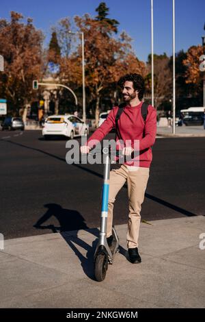 Glücklicher junger Mann, der mit einem elektrischen Roller auf der Straße steht Stockfoto