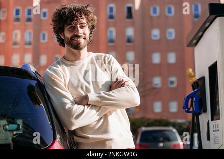 Lächelnder junger Mann, der an sonnigen Tagen neben einem Elektroauto steht Stockfoto