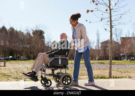 Eine Frau, die einem älteren Mann im Rollstuhl im Park assistiert Stockfoto