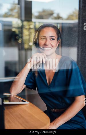 Lächelnde, aufmerksame Geschäftsfrau, die ein Headset trägt und durch Glas gesehen wird Stockfoto