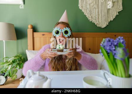 Fröhliche Frau mit Sonnenblumen-Sonnenbrille, Halteteller mit Cupcake im Bett zu Hause Stockfoto