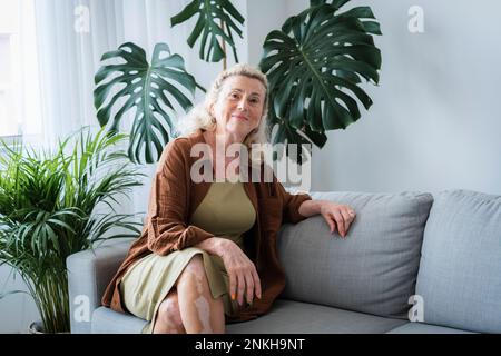 Lächelnde ältere Frau, die zu Hause auf dem Sofa sitzt Stockfoto