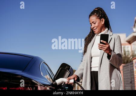 Eine lächelnde Frau steht mit einem Smartphone und lädt das Auto an sonnigen Tagen auf Stockfoto