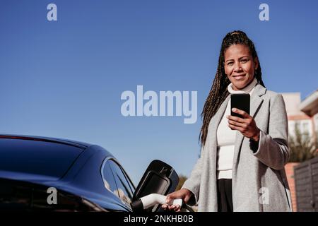 Lächelnde, reife Frau steht mit Smartphone und lädt das Auto an sonnigen Tagen auf Stockfoto