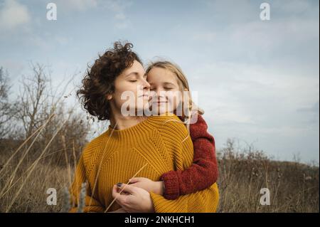 Lächelndes Mädchen, das Mutter von hinten umarmt, vor dem Himmel Stockfoto