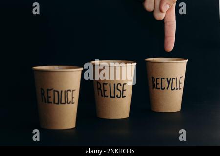 Die Hand eines Mannes zeigt auf einen wiederverwendbaren Becher mit Recycling-Text auf schwarzem Hintergrund Stockfoto