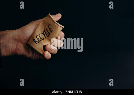 Hand eines Mannes, der einen wiederverwendbaren Becher vor schwarzem Hintergrund zerkleinert Stockfoto