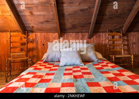 Queen-Size-Bett mit farbenfroher rot-blauer Steppdecke, antiken Holzstühlen im Sommer Master Schlafzimmer im oberen Stockwerk im alten 1840-Haus. Stockfoto