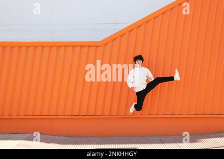 Ein sorgenloser junger Mann, der vor der orangefarbenen Wand auf einem Fußweg sprang Stockfoto