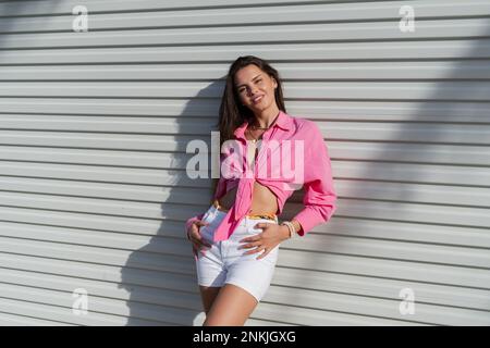Junge schöne Brünette Frau in einem rosa Hemd und weißen Denim-Shorts vor dem Hintergrund eines hellen Garagentürzaun Stockfoto