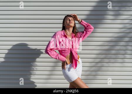 Junge schöne Brünette Frau in einem rosa Hemd und weißen Denim-Shorts vor dem Hintergrund eines hellen Garagentürzaun Stockfoto