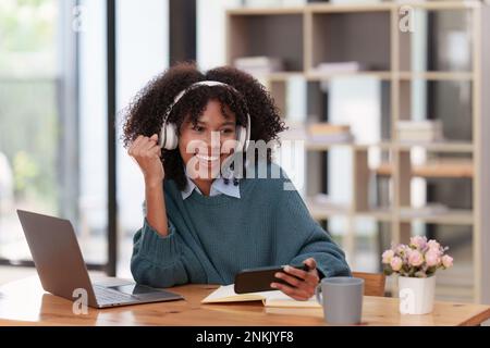 Aufgeregte junge schwarze afrikanische Frau trägt Kopfhörer und genießt Spiele über die App auf dem Mobiltelefon. Stockfoto