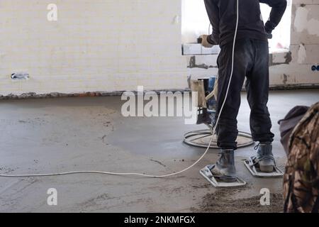 Rampfen und Schleifen von halbtrockenem Boden, der von einer Maschine mit rotierender Scheibe zum Nivellieren bearbeitet wird. Bau eines Betonfußbodens im Haus, ein Meister Stockfoto