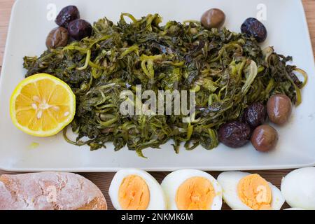 Gekochtes Wildgrün, Löwenzahn, Zitronen und natives Olivenöl extra auf einem Holztisch. Horta oder Wild Greens. Griechische Küche. Stockfoto