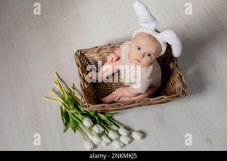 Das Baby in einem weißen Body mit Kaninchenohren auf dem Kopf sitzt in einem Korb mit einem Strauß Tulpen auf dem Boden. Hochwertiges Foto Stockfoto