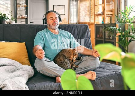 Reifer, übergewichtiger Mann mittleren Alters in kabellosen Kopfhörern, der sich zu Hause mit seiner Katze entspannt und Meditation führt, während er entspannte Musik auf dem Smartphone hört Stockfoto
