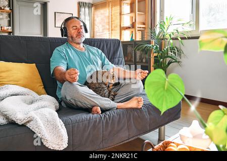 Reifer, übergewichtiger Mann mittleren Alters in kabellosen Kopfhörern, der sich zu Hause mit seiner Katze entspannt und Meditation führt, während er entspannte Musik auf dem Smartphone hört Stockfoto