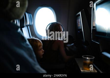 Ein Kind sitzt am Fenster im Flugzeug und schaut mit Kopfhörern heraus Stockfoto