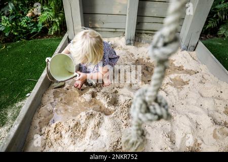 Ein Mädchen, das im Sandkasten mit einem Eimer Wasser spielt Stockfoto