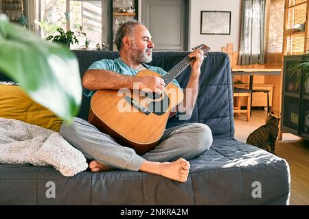 Ein bärtiger Mann mittleren Alters, der Akustikgitarre spielt, während er auf dem Sofa im hellen Wohnzimmer sitzt. Stockfoto