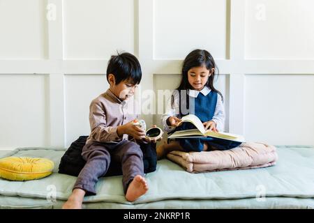 Zwei Kinder sitzen auf Kissen, Mädchen lesen Buch, Junge hält ein Objekt Stockfoto